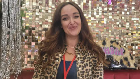 Laura Mears-Reynolds is sitting on a red sofa in front of a mirrored wall. She is wearing a leopard-print jacket and has long, brown hair. She is smiling at the camera.