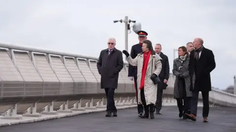 PA Media The Princess Royal walks across the Gull Bridge in Lowestoft, accompanied by local dignitaries, who are all smartly dressed