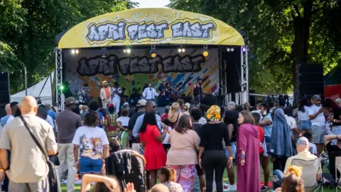 Mary Doggett/Afri-Fest East A festival stage in a park with a yellow banner with Afri-Fest East written in a graffiti style. Artists are performing on the stage and a crowd gathered in front.