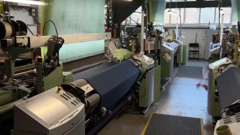 Richard Daniel/BBC A room full of machinery, including two green looms that have navy silk on them. In the far right, at the end of the room, is a white window.