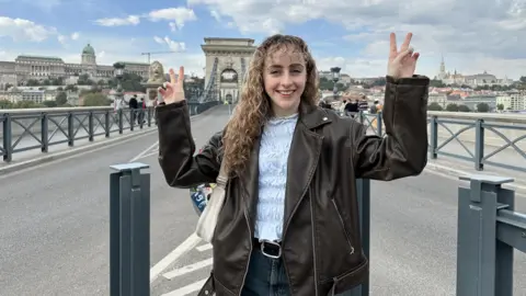 Marianne Ward A young woman with curly, dirty blonde hair smiles as she stands on a bridge in a European city. She is making two peace signs with her hands and is wearing a brown leather jacket, black denim jeans with a belt and a ruffled white top.