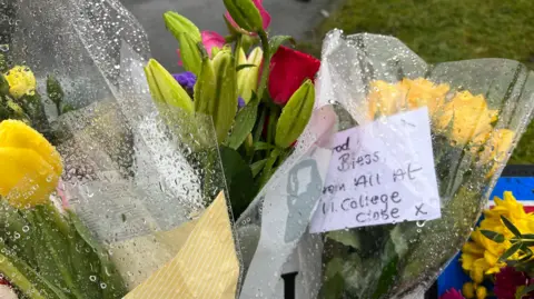 Oli Constable/BBC Several flower bouquets, including yellow tulips and roses, left at the scene where Chris Marriott died. One note, slightly soaked by raindrops, reads: "God bless, from all at 11 College Close x".
