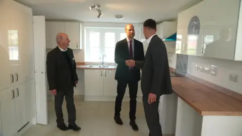 Prince William shaking hands with a man who is stood in a newly built kitchen. The doors and cupboards are white and the worktops are a light brown wood. There is also a man stood to the left of Prince William smiling at him. They are all wearing black suits.
