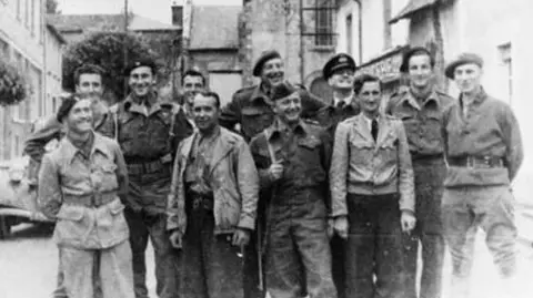 Imperial War Museum 11 resistance fighters in irregular uniform, smile for the camera ahead of battle in 1944. In the background is the wing of a vintage French car and rural buildings. 