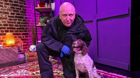 A picture of a dog handler next to a brown and white dog. They are in a studio with items surrounding them.