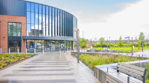 An wide paved road has a seat to one side and leads to a modern entrance of a railway station. It is a curved structure with long windows sitting above the doors and vegetation and grass on either side. A row of Victorian housing sits behind the station 