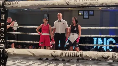 Bravehearts Amateur Boxing Club Dos boxeadores femeninos, en un kit rojo a la izquierda y negro a la derecha, paran a ambos lados de un árbitro de boxeo con camisa blanca y corbata de lazo negro, en el anillo de boxeo al final del combate.