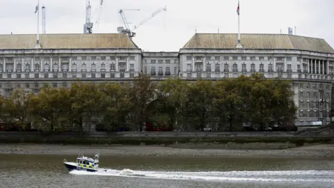 Reuters A view across the river Thames of MI5 headquarters, an imposing neo-classical building in grey stone. Flags are flying from the roof and construction cranes can be seen behind it. Part of the frontage is obscured by trees along the river, and a police boat is passing by on the water.