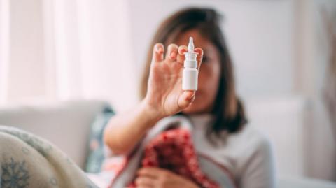 A nasal spray bottle in focus, a woman is holding it but she is out of focus behind it, she has shoulder length brown hair and is sat down with a blanker over her like she is recovering from an illness
