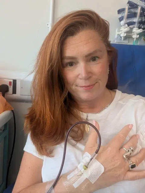 Sian Chathyoka Selfie of Sian sat in a blue hospital chair with a drip going into her hand. She is wearing a white t-shirt and has straight ginger hair. 