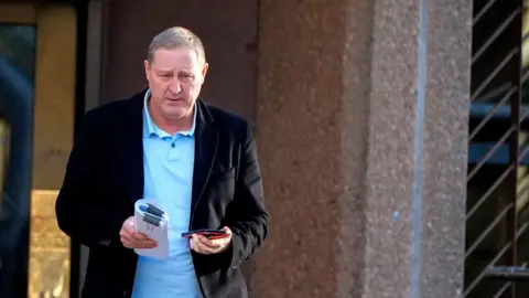 PA Media Paul Owen, with a black blazer over a blue shirt, walks away from Liverpool Crown Court holding a phone and a bundle of papers