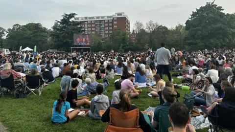 CJ Events A crowd of hundreds of people sitting on a large area of grass. They are sitting on the floor and on camping chairs. They are watching a large screen. There is a large building and green trees beyond the screen