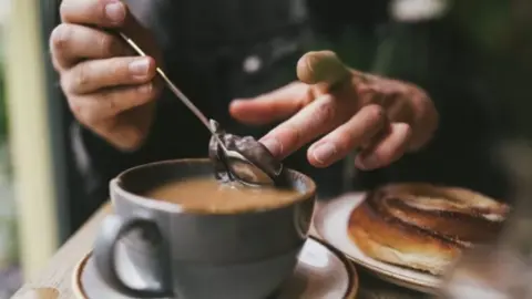 A cup of tea with a persons hands holding a spoon and taking the teabag out of the cup