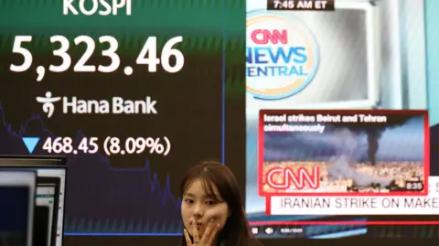 A currency dealer works in front of an electronic board displaying the Korea Composite Stock Price Index (KOSPI) and a news report on the U.S.-Israeli conflict with Iran, at the dealing room of a bank, in Seoul, South Korea, March 4, 2026.