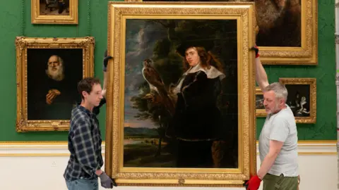 Royal Collection Trust Conservators rehanging a large framed portrait of a man with a bird of prey in Buckingham Palace's Picture Gallery, with other paintings displayed against emerald-green wall coverings.