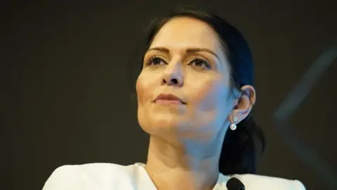 A headshot of Priti Patel as she looks above the camera. She wears a white blazer