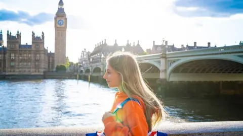 A young woman wearing a bright orange jacket and a Union Jack-coloured bag on her shoulder walks along the bank of the river Thames. Big Ben and the Houses of Parliament is in the background behind her.