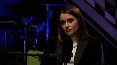 BBC News presenter Anne McAlpine has short brown hair and is wearing a black blazer. She is sitting in front of a microphone.