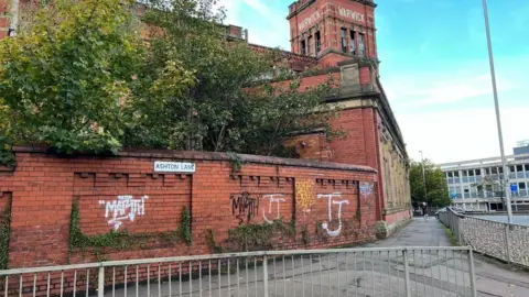 LDRS A wall by the mill covered in graffiti. The tower on the mill reads Warwick in white lettering. There is grey railing in front of the wall - which also has a sign which reads Ashton Lane.