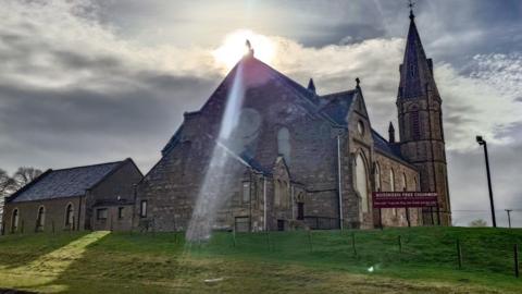 The Sun's beams hit the roof of a church and highlight part of the side. The sky is cloudy