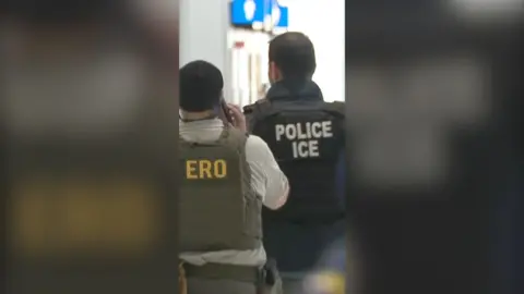 The backs of two ICE agents one in green wearing bullet proof vests