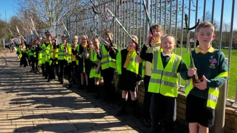 A line of year three primary school pupils wearing hi-vis vests cheering and waving litter pickers in the air. 