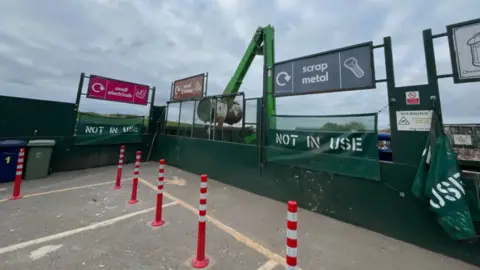 The recycling centre. There are bollards, alongside a pathway. There are small gaps in a green fence where residents can drop their waste through. There is a crane on the other side of the fencing.
