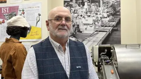 A man with a shave head and grey beard stands in a museum in front of factory equipment and memorabilia, including a silver wrapping machine with an orange wrapped sweet on a chute. He is wearing glasses and a blue checked waistcoat over a light checked shirt.