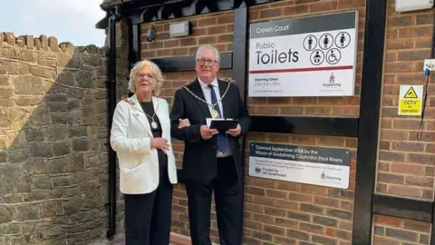 The Mayor and Mayor Consort dressed in their ceremonial chains stand outside the toilets made out of red bricks. A plaque on the toilet wall reads "Opened September 2024 by the Mayor of Godalming councillor Paul Rivers"