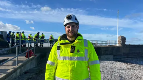 Drew Parkinson smiling towards the camera and stood on Brixham beach. He is wearing a yellow hi-vis jacket and white hat. There are recruits in the background.