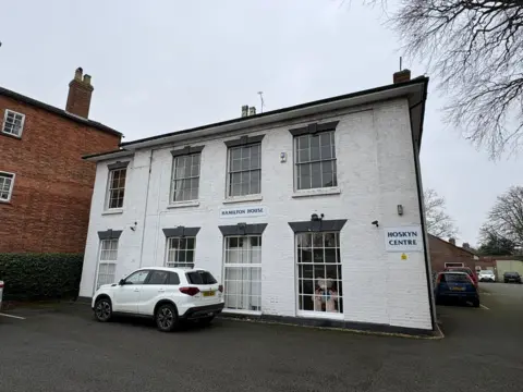 A white car is parked in front of a white two-story building with eight large windows. There is a sign on the building which says Hoskyn Centre