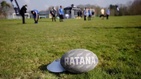 The Matt Ratana Foundation A rugby ball with RATANA printed on it with rugby players standing and stretching in the background.
