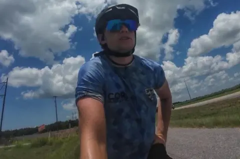 Joe Dick A man in a light blue England football shirt, cycle helmet and wrap around sunglasses sits on a bike on the side of rural road.
