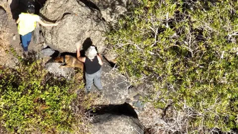 Handout Aerial image of two women and a dog searching among rocks and scrub