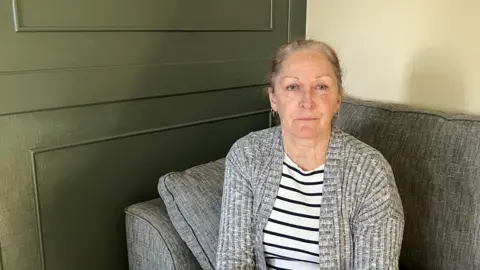 Casey Tremain sits on a sofa and is looking at the camera. Her brown/grey hair is pulled back and she is wearing a grey cardigan and black and white striped top.