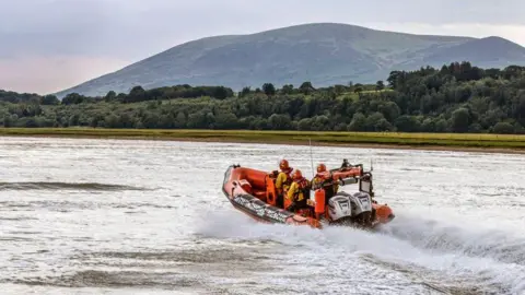A lifeboat skipping across the water surface on the Solway estuary with trees and hills in the background