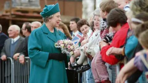 Getty Images Queen Elizabeth II who is wearing a green/blue coat and hat. She's talking to people in the crowd and is holding pink, white and purple flowers.