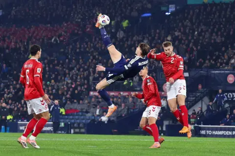 Getty Images Scott McTominay rises to execute an overhead kick goal amid an aerial challenge from a Danish defender. He is wearing a full navy blue Scotland kit. Two Danish players wearing red tops, white shorts and red socks look on.