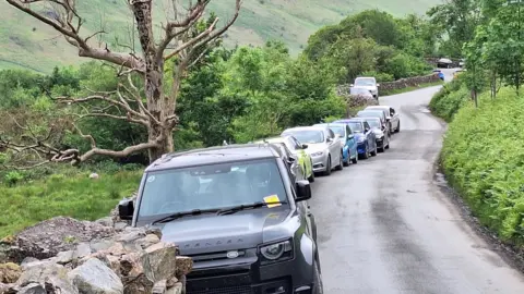 A row of vehicles parked along a narrow country road. The Land Rover at the front of the line has a yellow parking ticket stuck to its front window.