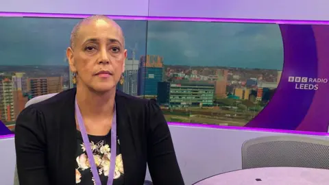 A woman with a shaved head and wearing a black cardigan and dress with a white floral pattern sitting in a radio studio with a purple background