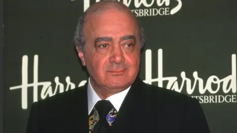 BBC Mohamed Al Fayed wears a dark suit and stands in front of a dark green sign which reads 'Harrods, Kinightsbridge'.