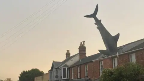 The Headington Shark sculpture - the body of a shark Diving into the roof of a house -  with other terraced properties on either side. The sky is a dusky blue/grey colour with a hue of sunset yellows and golds to the left of the image.