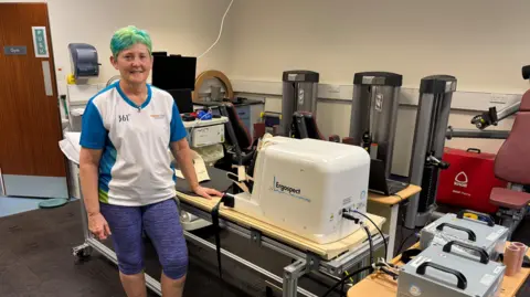 BBC Julie Smith, with died green hair, wearing a white and blue tee-shirt and purple/black pattered leggings to just below the knee stands to camera left with her hand resting on a horizontal stepping machine. Fitness and monitoring instruments of different kind are visible around the room