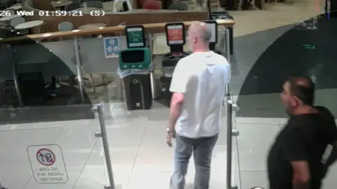 North Yorkshire Police A man in a white t-shirt and grey jeans and shaven head stands with his back to the CCTV camera. Behind him is a man in a black t-shirt with dark hair. In front of them are two bins and an empty seating area.