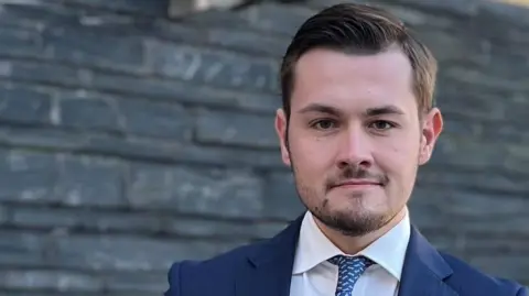 Joel Williams Joel Williams stood in front of the Senedd building wearing a blue suit, blue tie and white shirt.