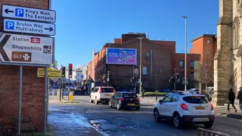 Three vehicles queuing at a red traffic light. There are road signs on the left of the image and buildings in the background. The picture was taken on a sunny day.