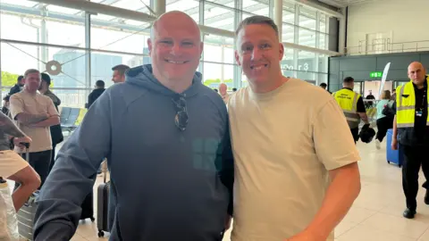 Davina Ramos/BBC Bob Firth and Nathan Hall stood together smiling at Manchester Airport. Bob has a bald head and us wearing a grey designer hoodie. Nathan has short brown hair and is wearing a white T-Shirt