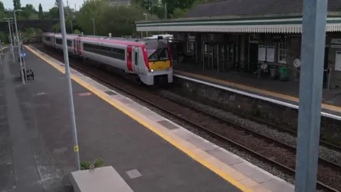 A passenger train is pulling into a small outdoor railway station with two platforms, with a stone station building and a few people waiting nearby. The tracks curve away under a bridge in the background, with signals showing red and trees and houses surrounding the area.