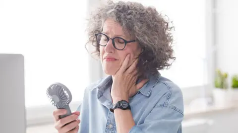 A woman with curly greyish hair, wearing blue-rimmed glasses and a blue denim shirt, holds a small portable fan in front of her face while closing her eyes and holding her hand to her cheek.