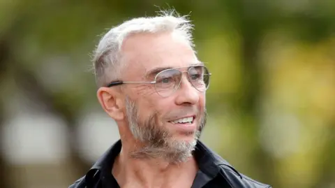 James Holder is seen in a head and shoulders image wearing a black shirt and metal-framed glasses. He has a beard and short hair which is a mixture of dark and grey